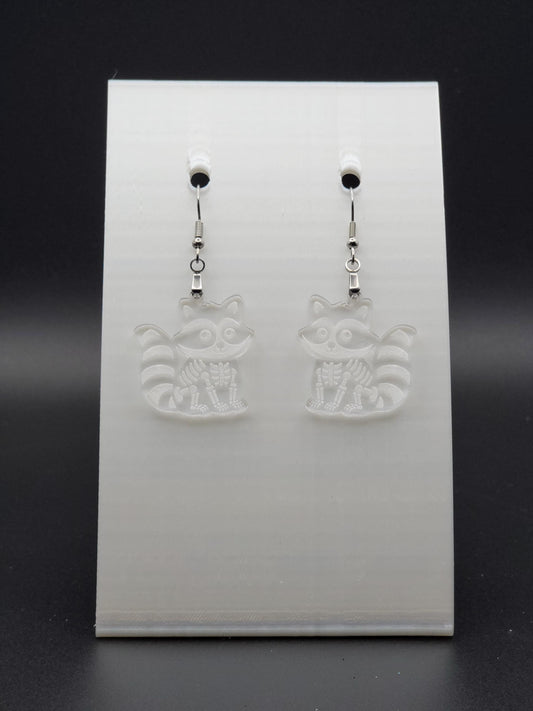 Clear acrylic skeleton raccoon dangle earring with engraved bone details and stainless steel hook, displayed on a white jewelry card. Laser-cut, handmade spooky jewelry.