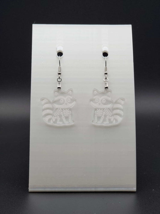 Clear acrylic skeleton raccoon dangle earring with engraved bone details and stainless steel hook, displayed on a white jewelry card. Laser-cut, handmade spooky jewelry.