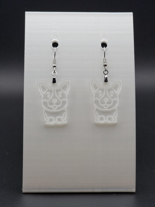 Pair of clear acrylic corgi earrings engraved with a playful face. Shown on a white earring card.
