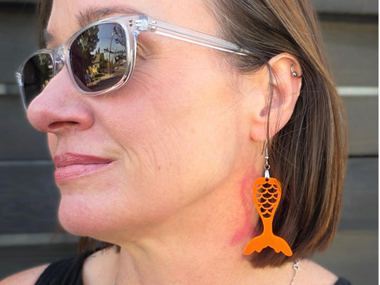 Close-up profile of a woman wearing a fluorescent acrylic mermaid tail earring outdoors, showing the earring's length and movement against her jaw and neck.