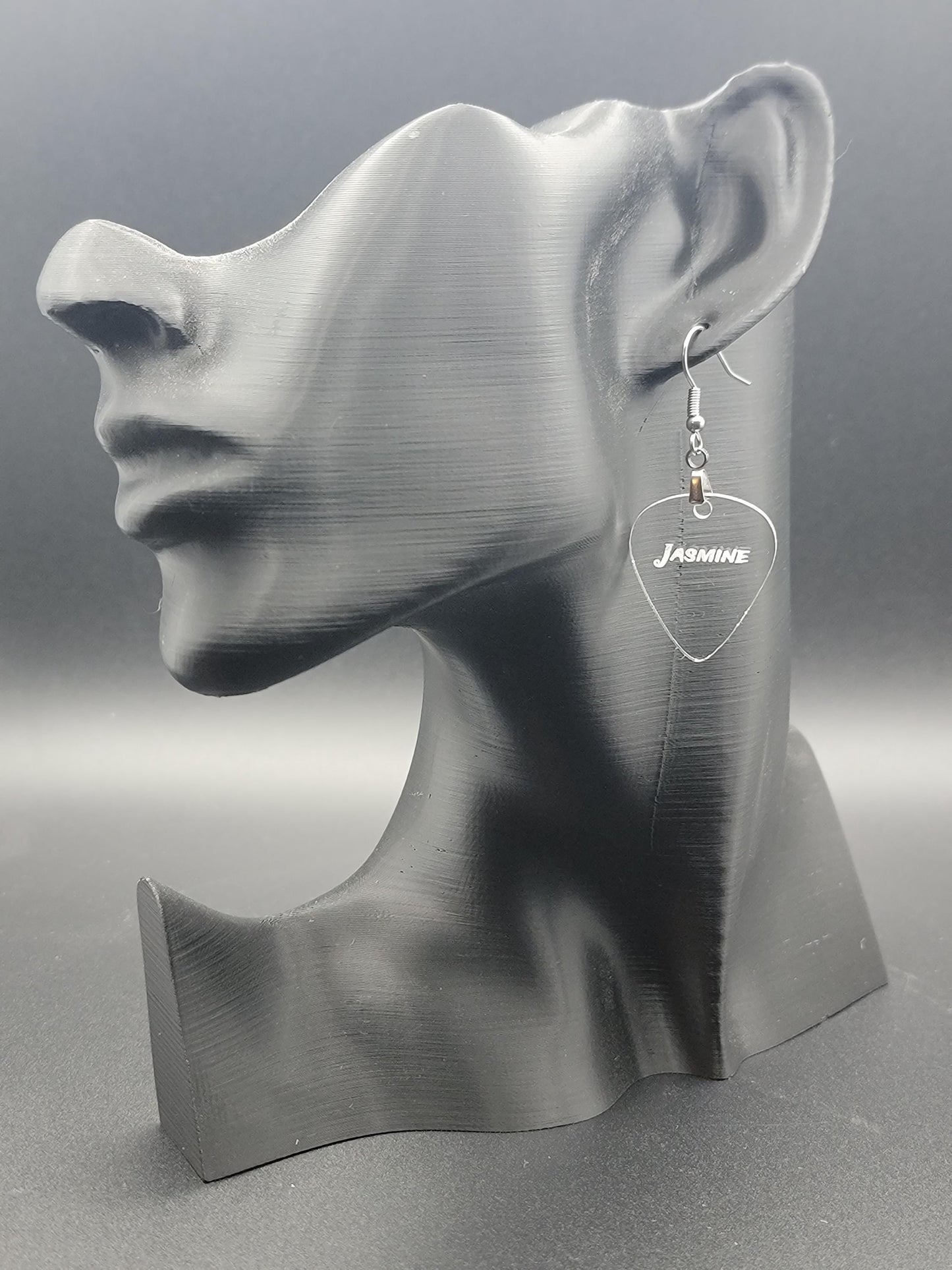 Clear acrylic guitar pick earring with "Jasmine" engraved, shown on a modern matte black mannequin head. Fun, personalized jewelry for music lovers.