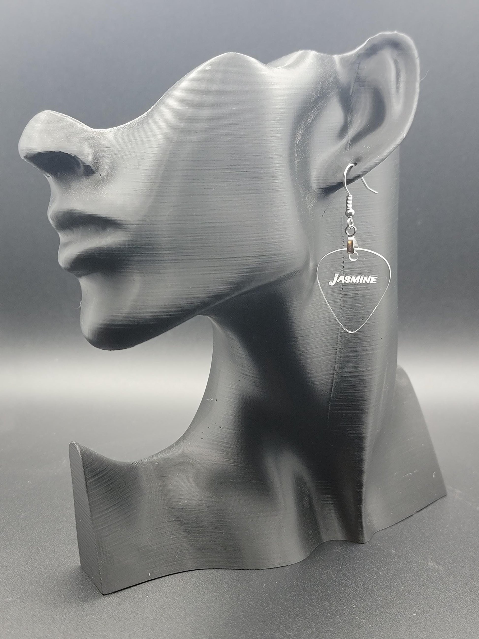 Clear acrylic guitar pick earring with "Jasmine" engraved, shown on a modern matte black mannequin head. Fun, personalized jewelry for music lovers.