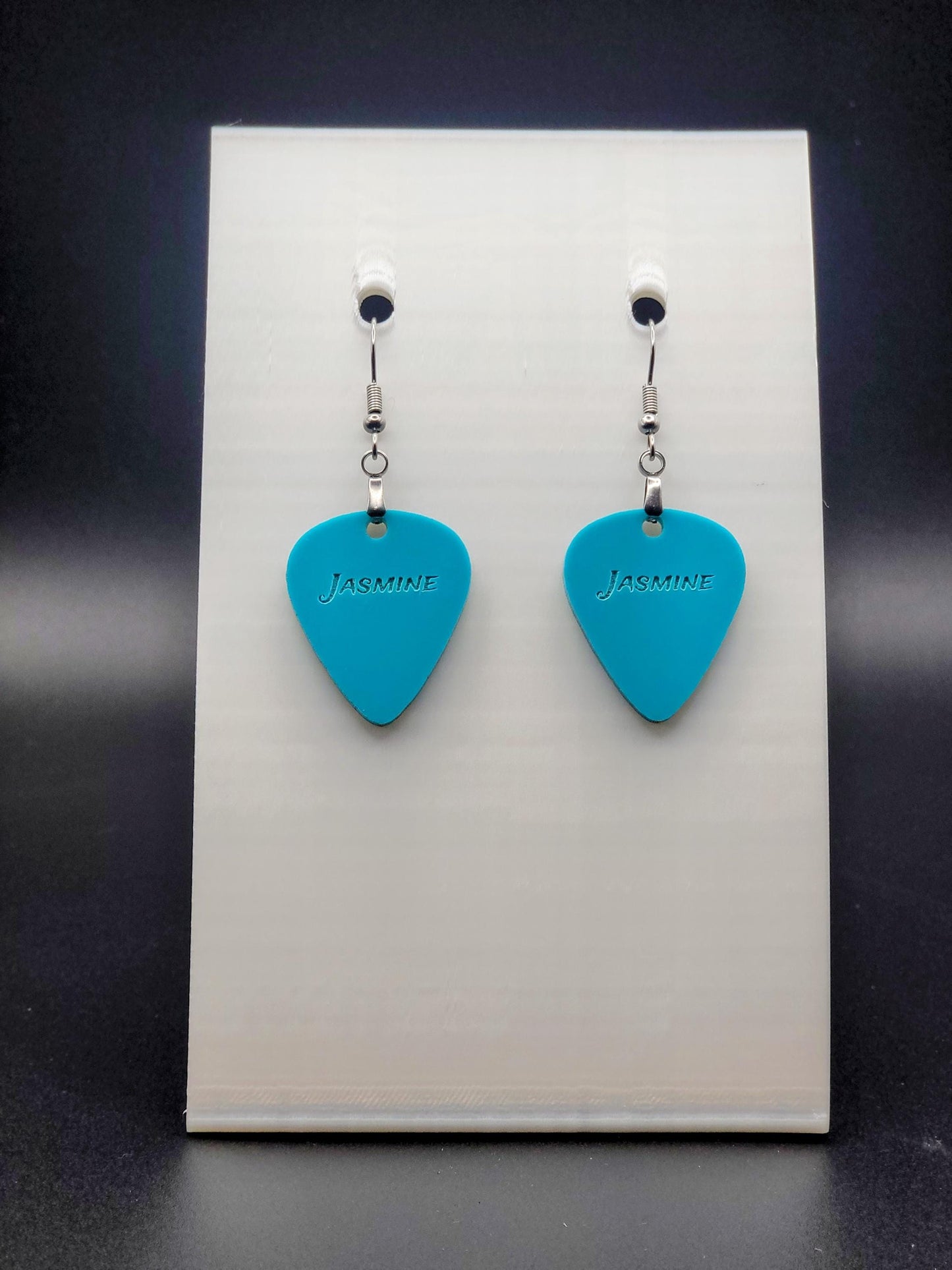 Teal acrylic guitar pick earring with "Jasmine" engraved, shown on a white jewelry card. Fun, personalized jewelry for music lovers.