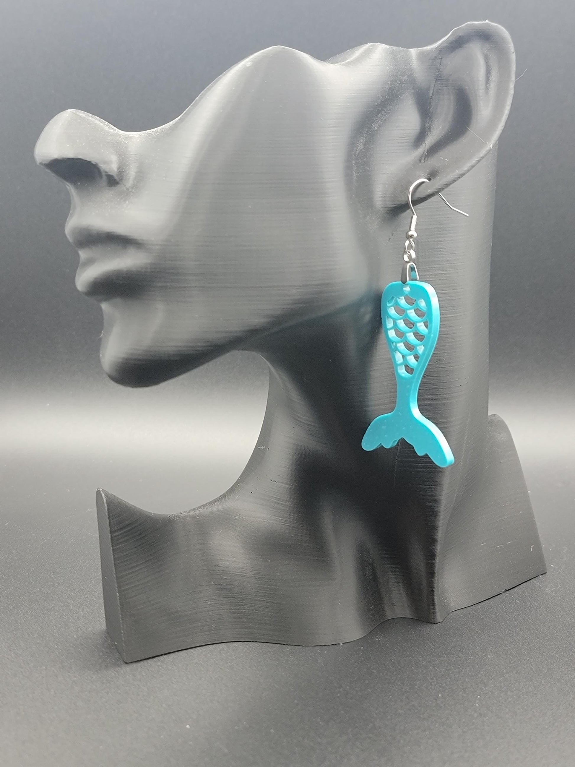 Teal acrylic mermaid tail earrings with engraved scale details and stainless steel hooks, shown on a modern matte black mannequin head. Glowforge laser-cut, handmade ocean-themed jewelry.