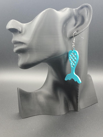 Teal acrylic mermaid tail earrings with engraved scale details and stainless steel hooks, shown on a modern matte black mannequin head. Glowforge laser-cut, handmade ocean-themed jewelry.