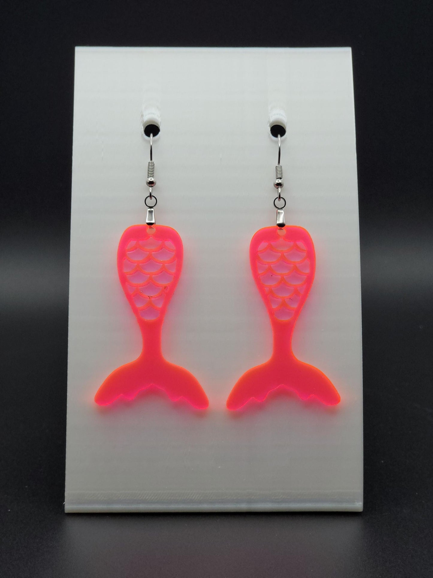 Fluorescent acrylic mermaid tail earrings with engraved scale details and stainless steel hooks, shown on a white jewelry card. Glowforge laser-cut, handmade ocean-themed jewelry.