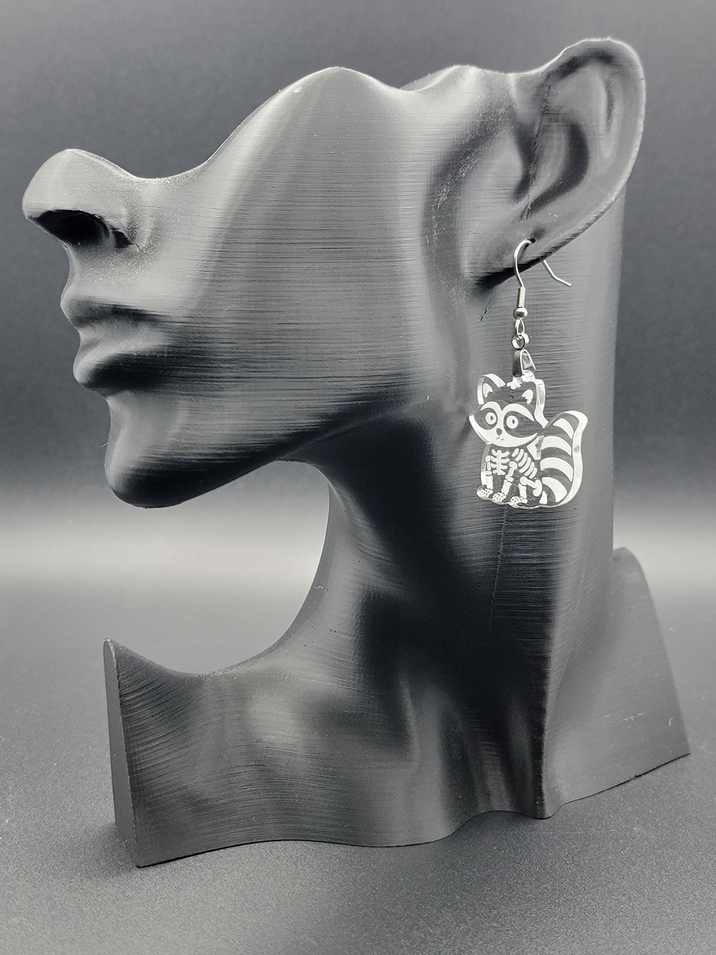 Clear acrylic skeleton raccoon dangle earring with engraved bone details and stainless steel hook, displayed on a matte black mannequin. Laser-cut, handmade spooky jewelry.