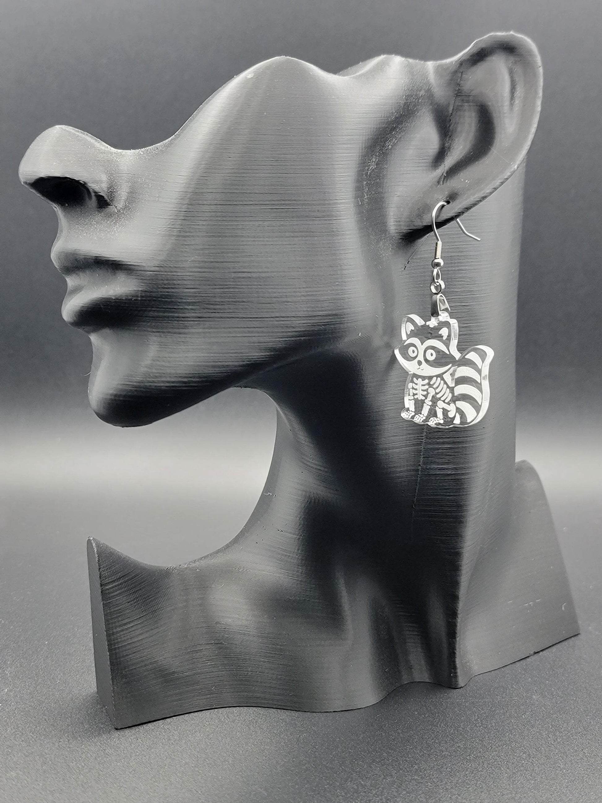 Clear acrylic skeleton raccoon dangle earring with engraved bone details and stainless steel hook, displayed on a matte black mannequin. Laser-cut, handmade spooky jewelry.