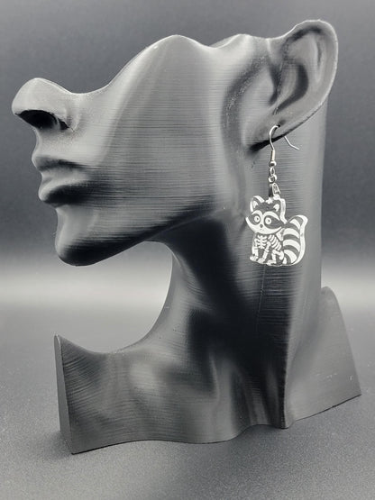 Clear acrylic skeleton raccoon dangle earring with engraved bone details and stainless steel hook, displayed on a matte black mannequin. Laser-cut, handmade spooky jewelry.