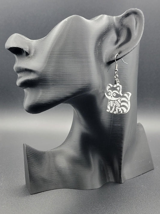 Clear acrylic skeleton raccoon dangle earring with engraved bone details and stainless steel hook, displayed on a matte black mannequin. Laser-cut, handmade spooky jewelry.