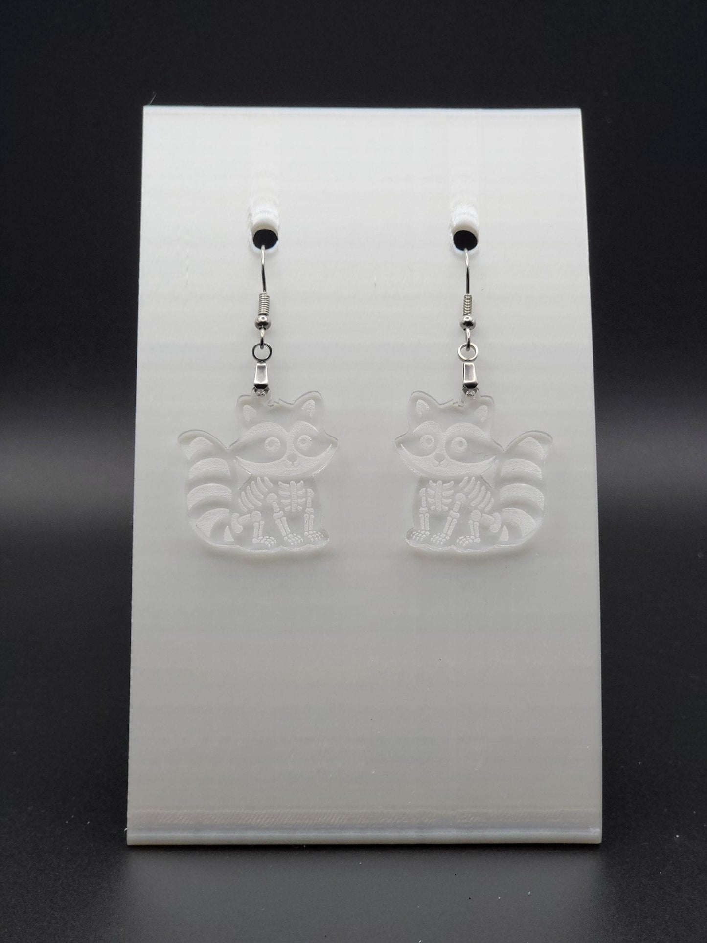 Clear acrylic skeleton raccoon dangle earring with engraved bone details and stainless steel hook, displayed on a white jewelry card. Laser-cut, handmade spooky jewelry.