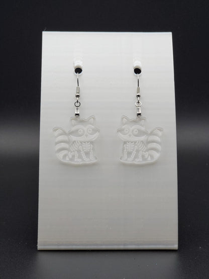 Clear acrylic skeleton raccoon dangle earring with engraved bone details and stainless steel hook, displayed on a white jewelry card. Laser-cut, handmade spooky jewelry.