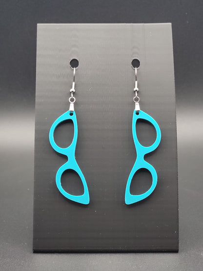 Teal acrylic cat-eye glasses dangle earrings on a matte black jewelry card. Handmade, laser-cut retro jewelry.
