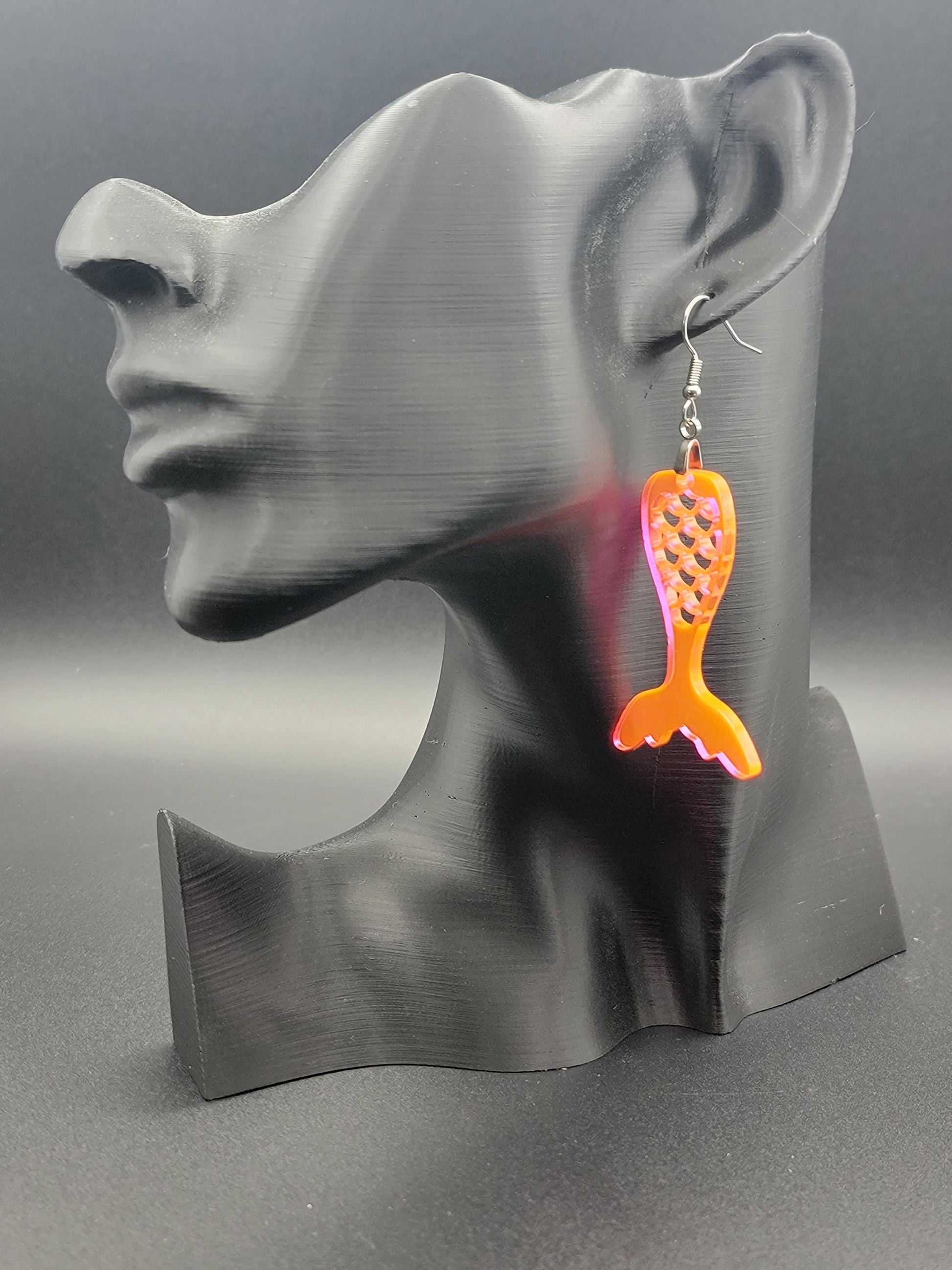 Fluorescent acrylic mermaid tail earrings with engraved scale details and stainless steel hooks, shown on a modern matte black mannequin head. Glowforge laser-cut, handmade ocean-themed jewelry.