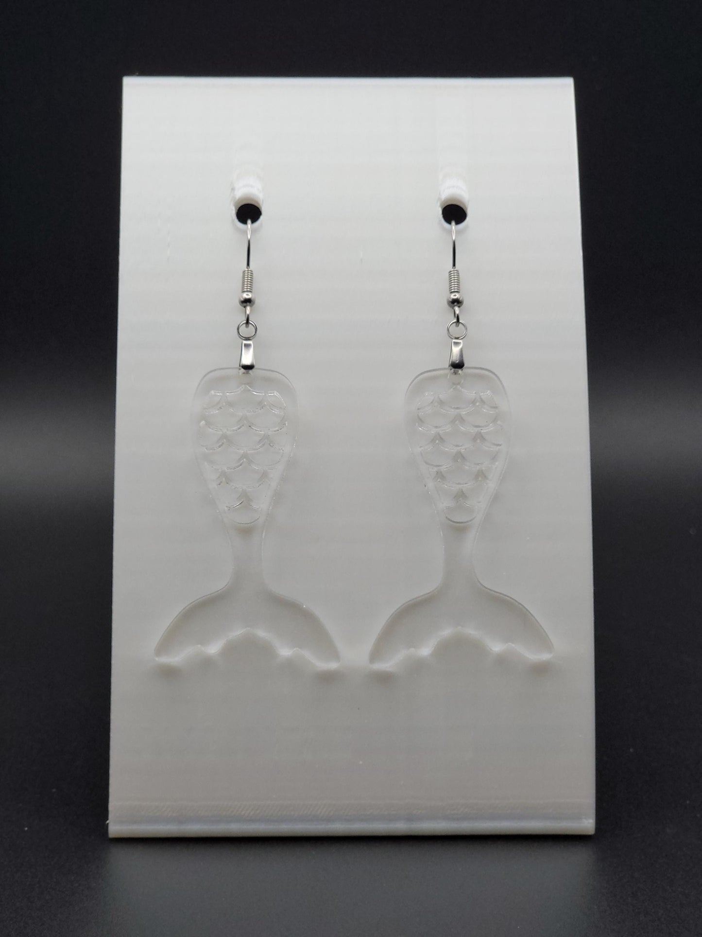 Clear acrylic mermaid tail earrings with engraved scale details and stainless steel hooks, shown on a white jewelry card. Glowforge laser-cut, handmade ocean-themed jewelry.