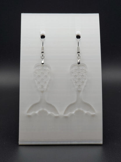 Clear acrylic mermaid tail earrings with engraved scale details and stainless steel hooks, shown on a white jewelry card. Glowforge laser-cut, handmade ocean-themed jewelry.