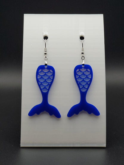 Blue acrylic mermaid tail earrings with engraved scale details and stainless steel hooks, shown on a white jewelry card. Glowforge laser-cut, handmade ocean-themed jewelry.