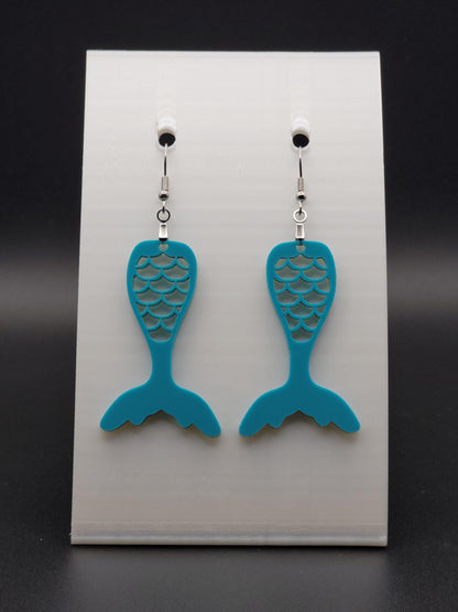 Teal acrylic mermaid tail earrings with engraved scale details and stainless steel hooks, shown on a white jewelry card. Glowforge laser-cut, handmade ocean-themed jewelry.