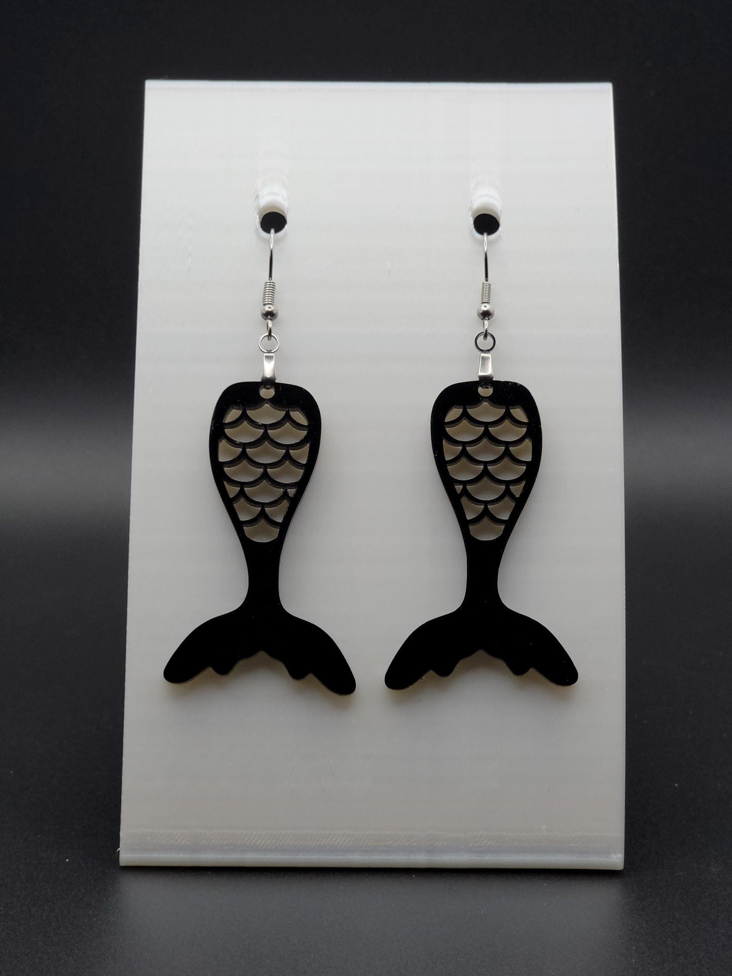 Black acrylic mermaid tail earrings with engraved scale details and stainless steel hooks, shown on a white jewelry card. Glowforge laser-cut, handmade ocean-themed jewelry.