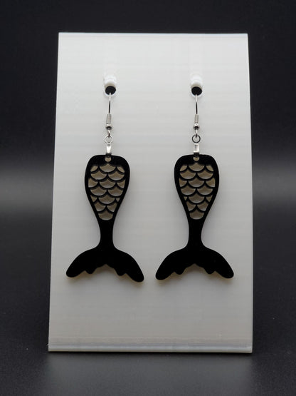Black acrylic mermaid tail earrings with engraved scale details and stainless steel hooks, shown on a white jewelry card. Glowforge laser-cut, handmade ocean-themed jewelry.
