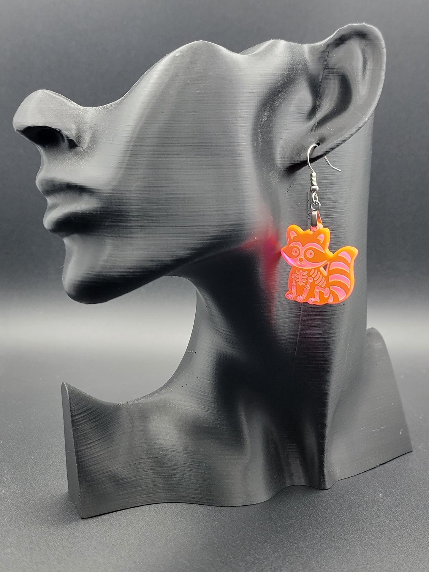 Fluorescent acrylic skeleton raccoon dangle earring with engraved bone details and stainless steel hook, displayed on a matte black mannequin. Laser-cut, handmade spooky jewelry.