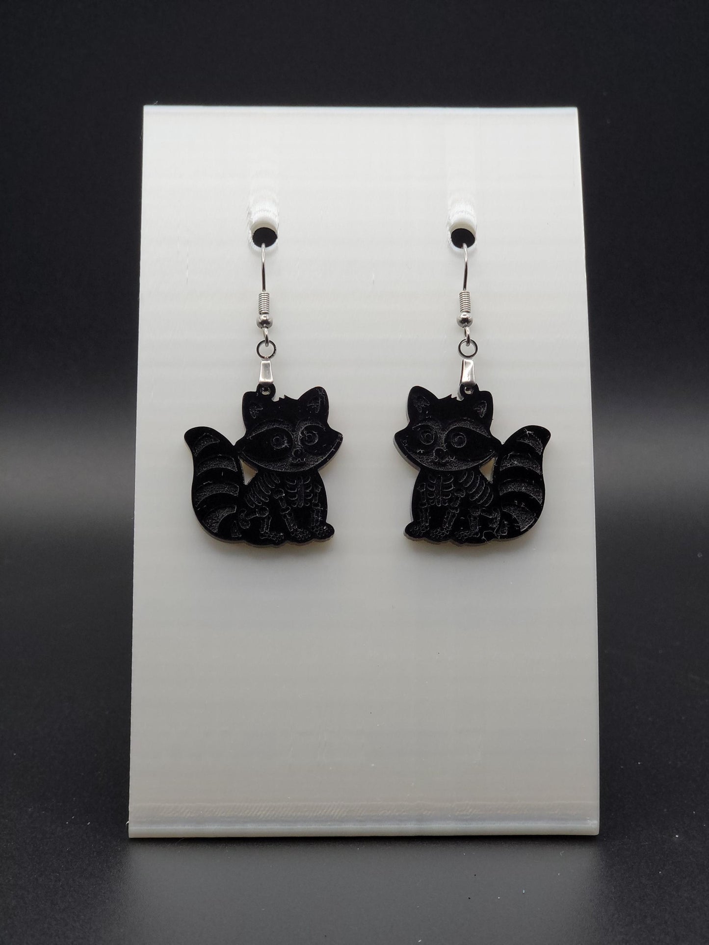 Black acrylic skeleton raccoon dangle earring with engraved bone details and stainless steel hook, displayed on a white jewelry card. Laser-cut, handmade spooky jewelry.