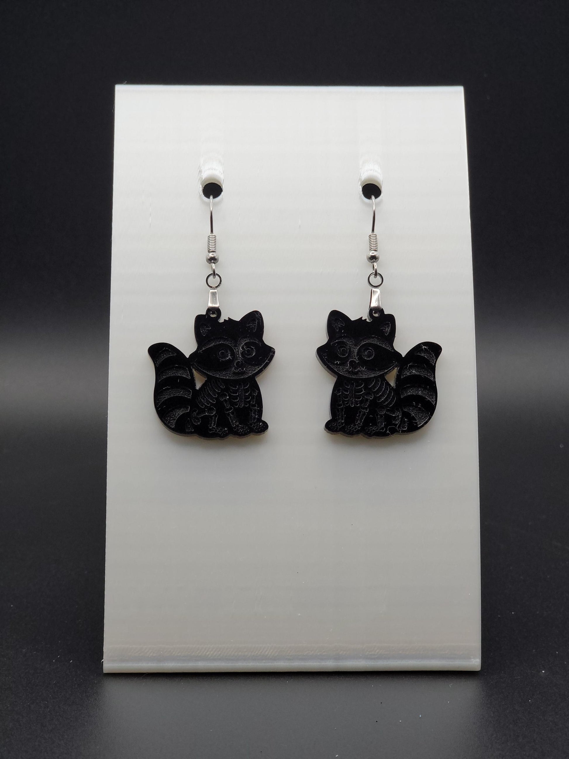 Black acrylic skeleton raccoon dangle earring with engraved bone details and stainless steel hook, displayed on a white jewelry card. Laser-cut, handmade spooky jewelry.