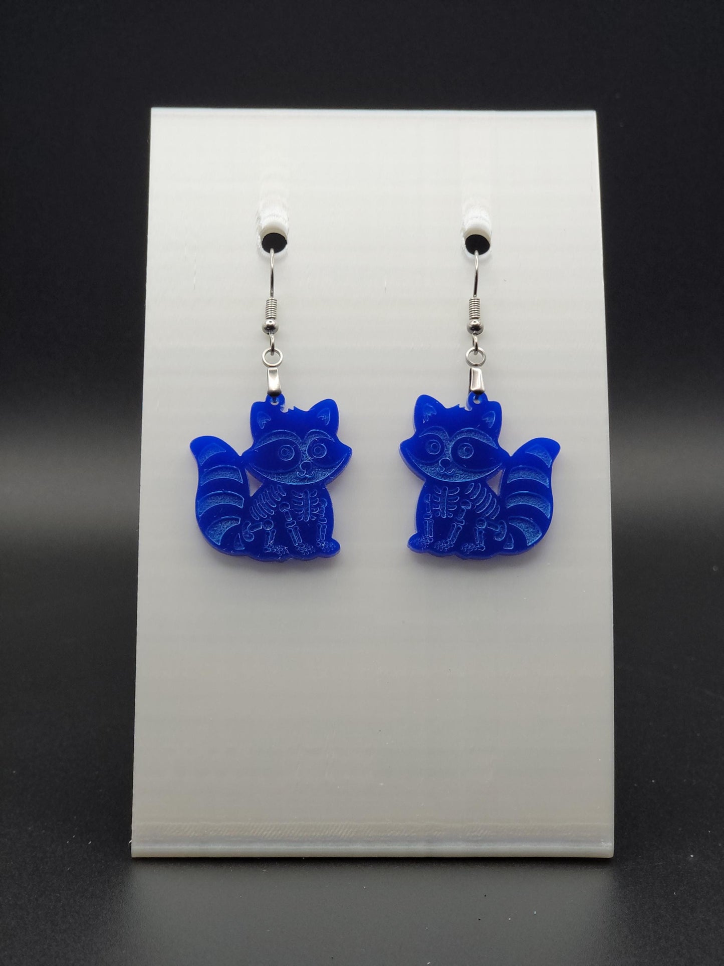 Blue acrylic skeleton raccoon dangle earring with engraved bone details and stainless steel hook, displayed on a white jewelry card. Laser-cut, handmade spooky jewelry.