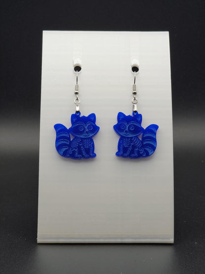Blue acrylic skeleton raccoon dangle earring with engraved bone details and stainless steel hook, displayed on a white jewelry card. Laser-cut, handmade spooky jewelry.