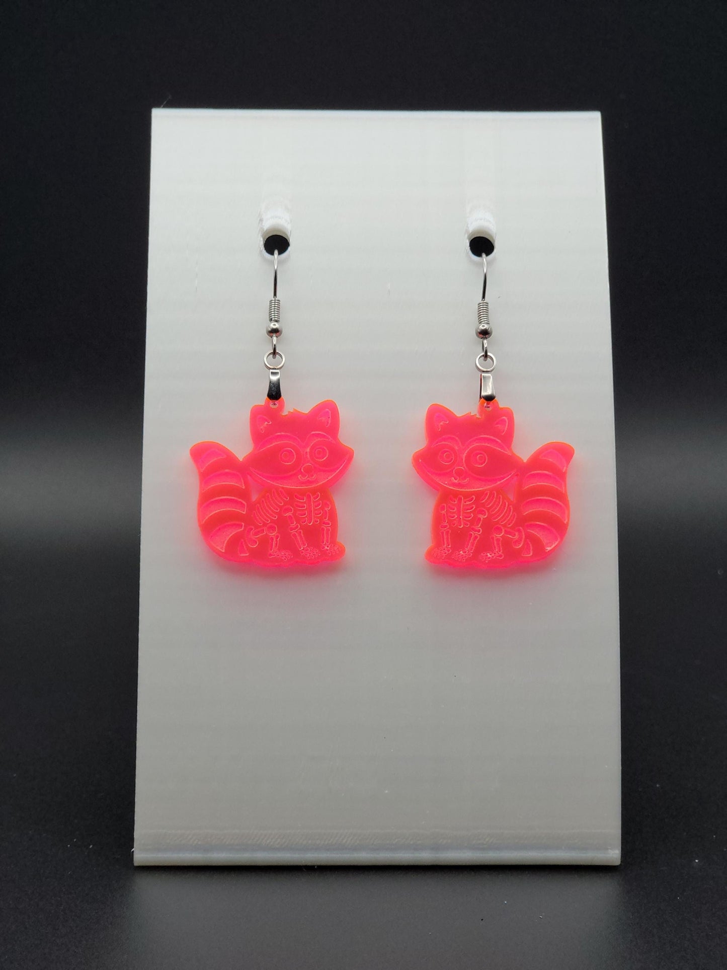 Fluorescent acrylic skeleton raccoon dangle earring with engraved bone details and stainless steel hook, displayed on a white jewelry card. Laser-cut, handmade spooky jewelry.