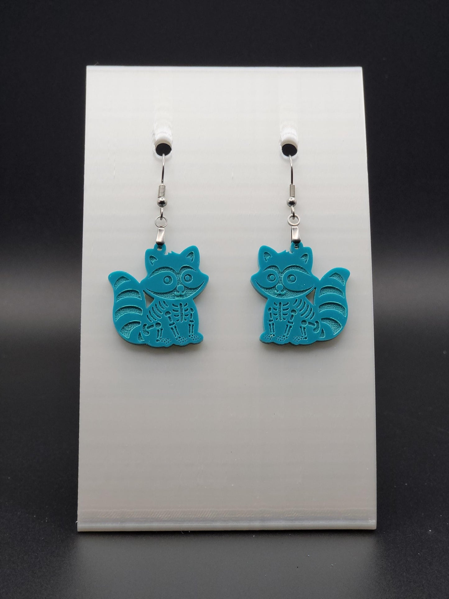 Teal acrylic skeleton raccoon dangle earring with engraved bone details and stainless steel hook, displayed on a white jewelry card. Laser-cut, handmade spooky jewelry.