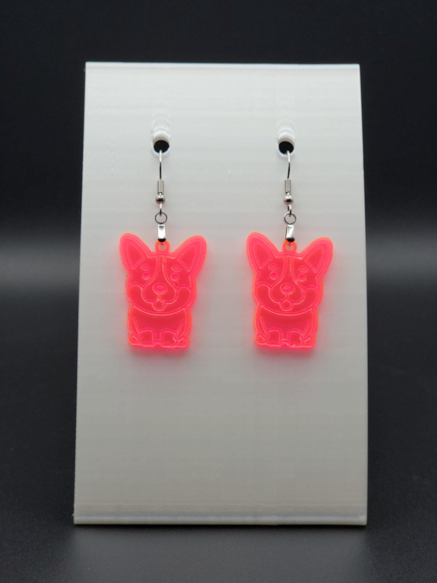 Pair of fluorescent acrylic corgi earrings engraved with a playful face. Shown on a white earring card.