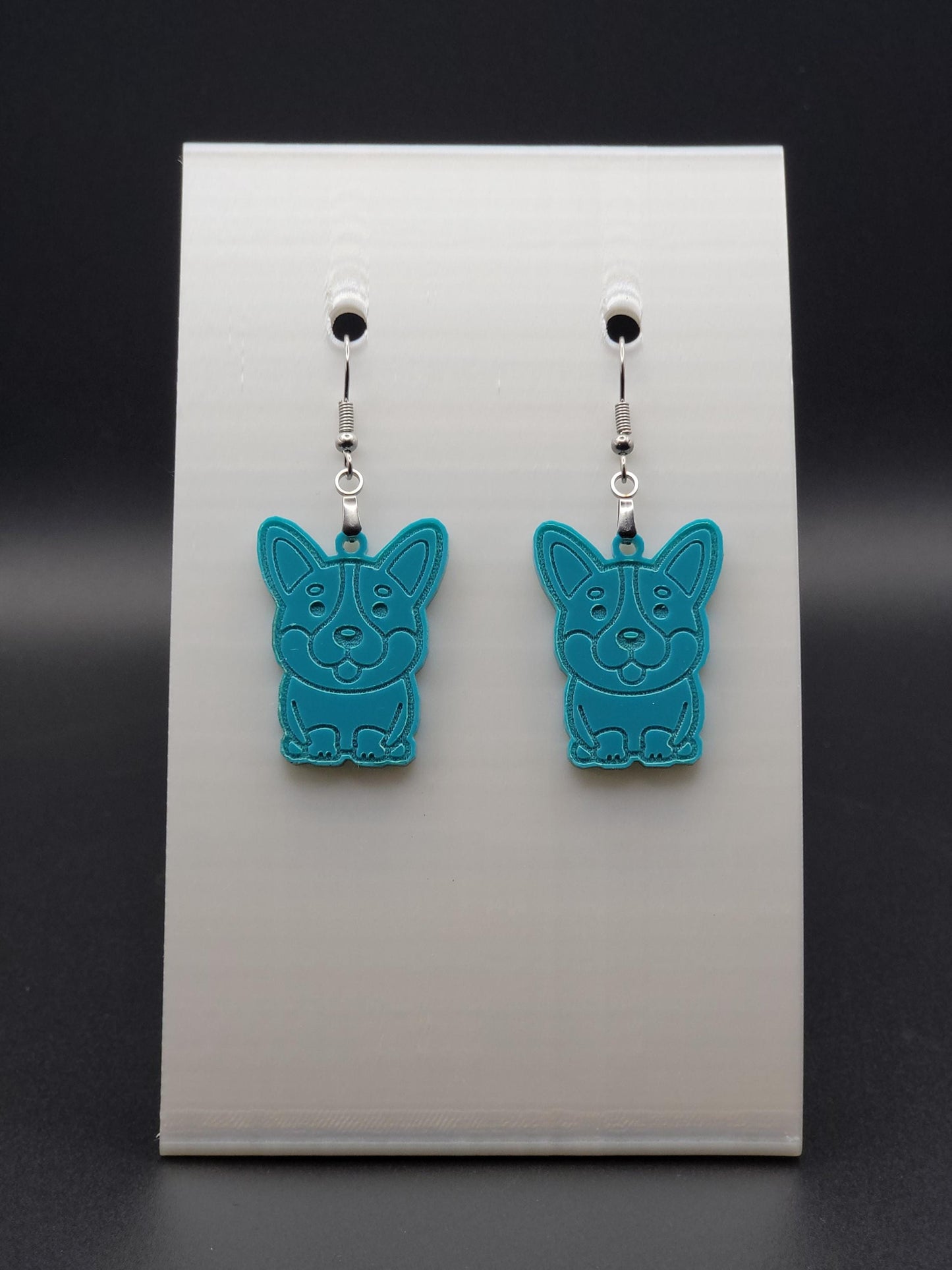 Pair of teal acrylic corgi earrings engraved with a playful face. Shown on a white earring card.