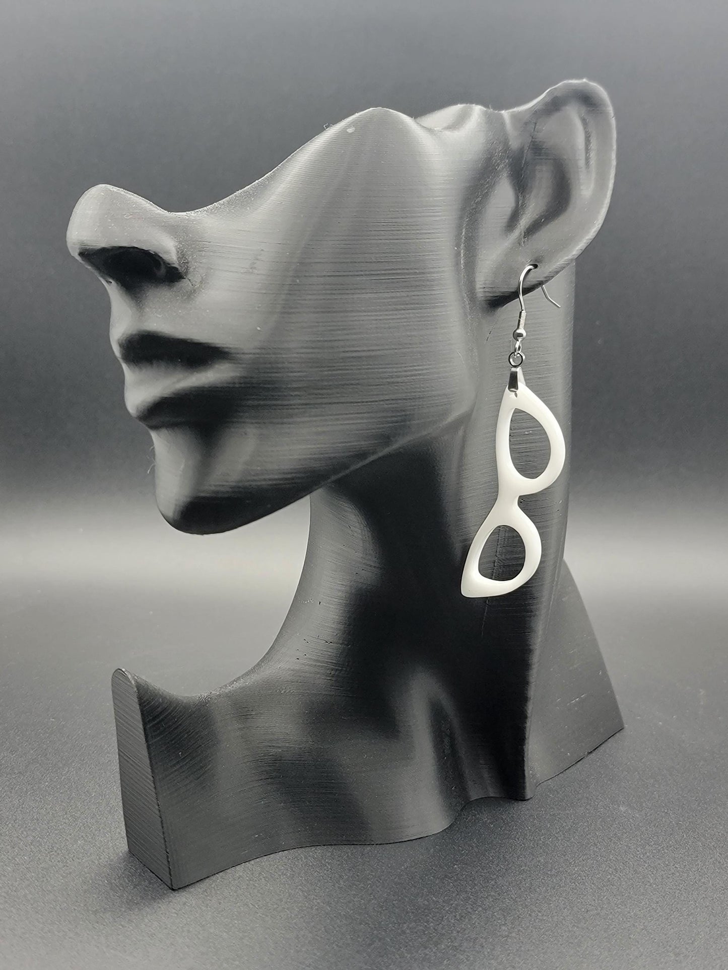 Single white dangle earring in the shape of cat-eye glasses on a black bust display, showcasing its bold, retro silhouette.