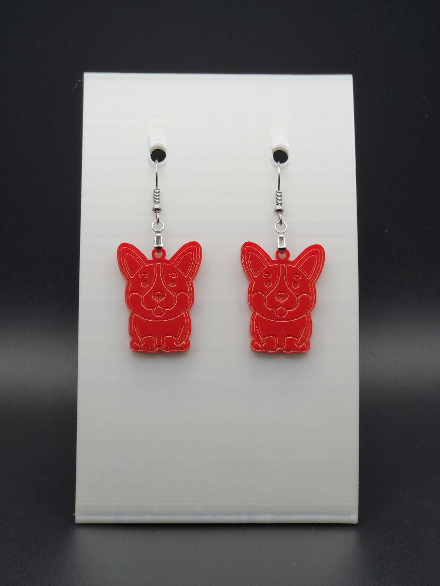 Pair of red acrylic corgi earrings engraved with a playful face. Shown on a black earring card.