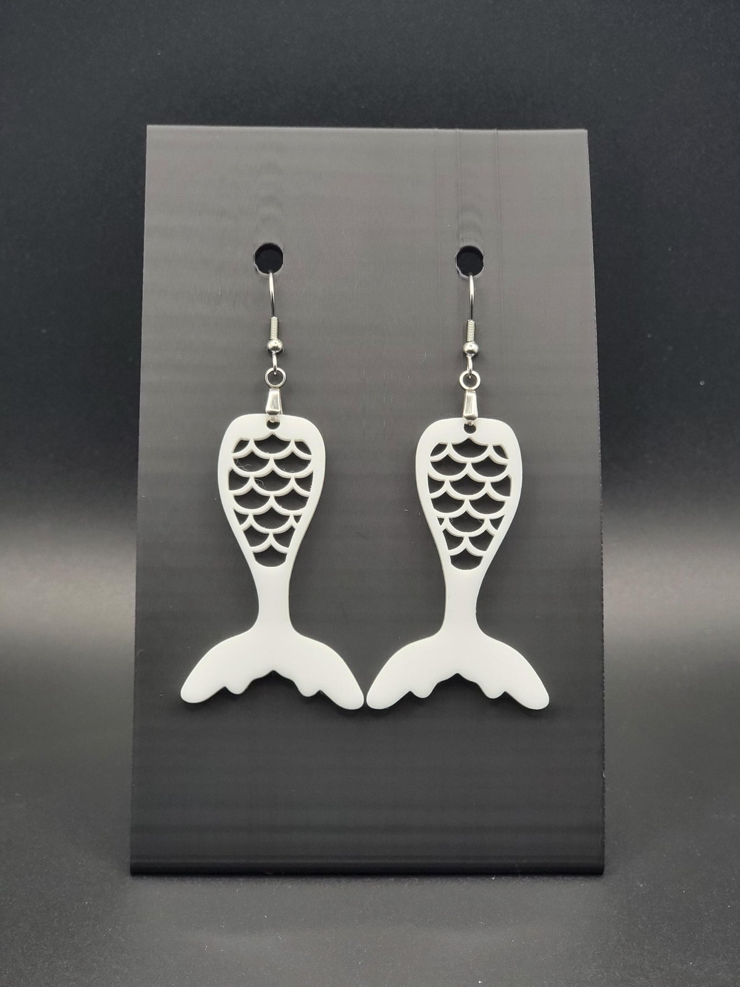 White acrylic mermaid tail earrings with engraved scale details and stainless steel hooks, shown on a black jewelry card. Glowforge laser-cut, handmade ocean-themed jewelry.