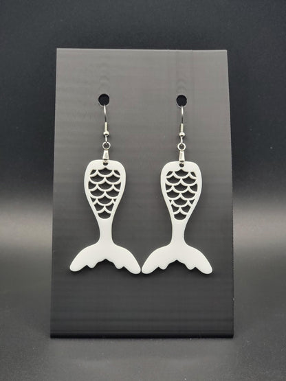 White acrylic mermaid tail earrings with engraved scale details and stainless steel hooks, shown on a black jewelry card. Glowforge laser-cut, handmade ocean-themed jewelry.