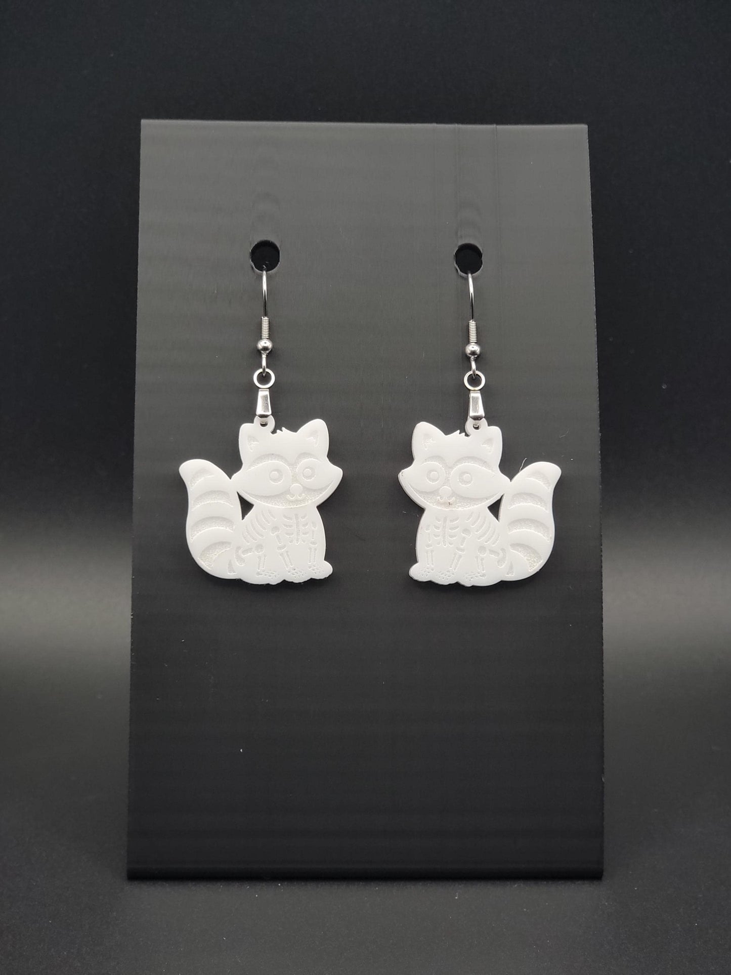 White acrylic skeleton raccoon dangle earring with engraved bone details and stainless steel hook, displayed on a black jewelry card. Laser-cut, handmade spooky jewelry.