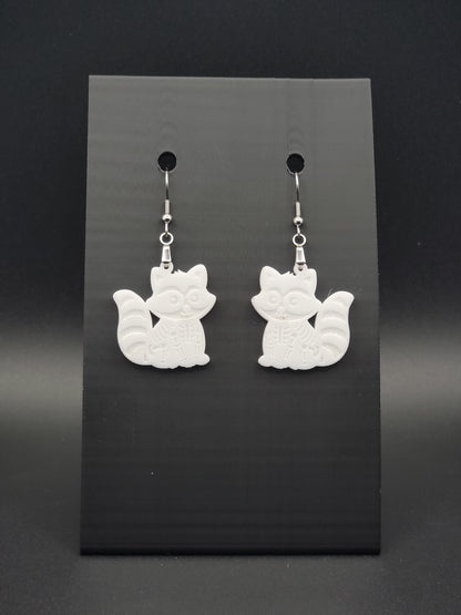 White acrylic skeleton raccoon dangle earring with engraved bone details and stainless steel hook, displayed on a black jewelry card. Laser-cut, handmade spooky jewelry.