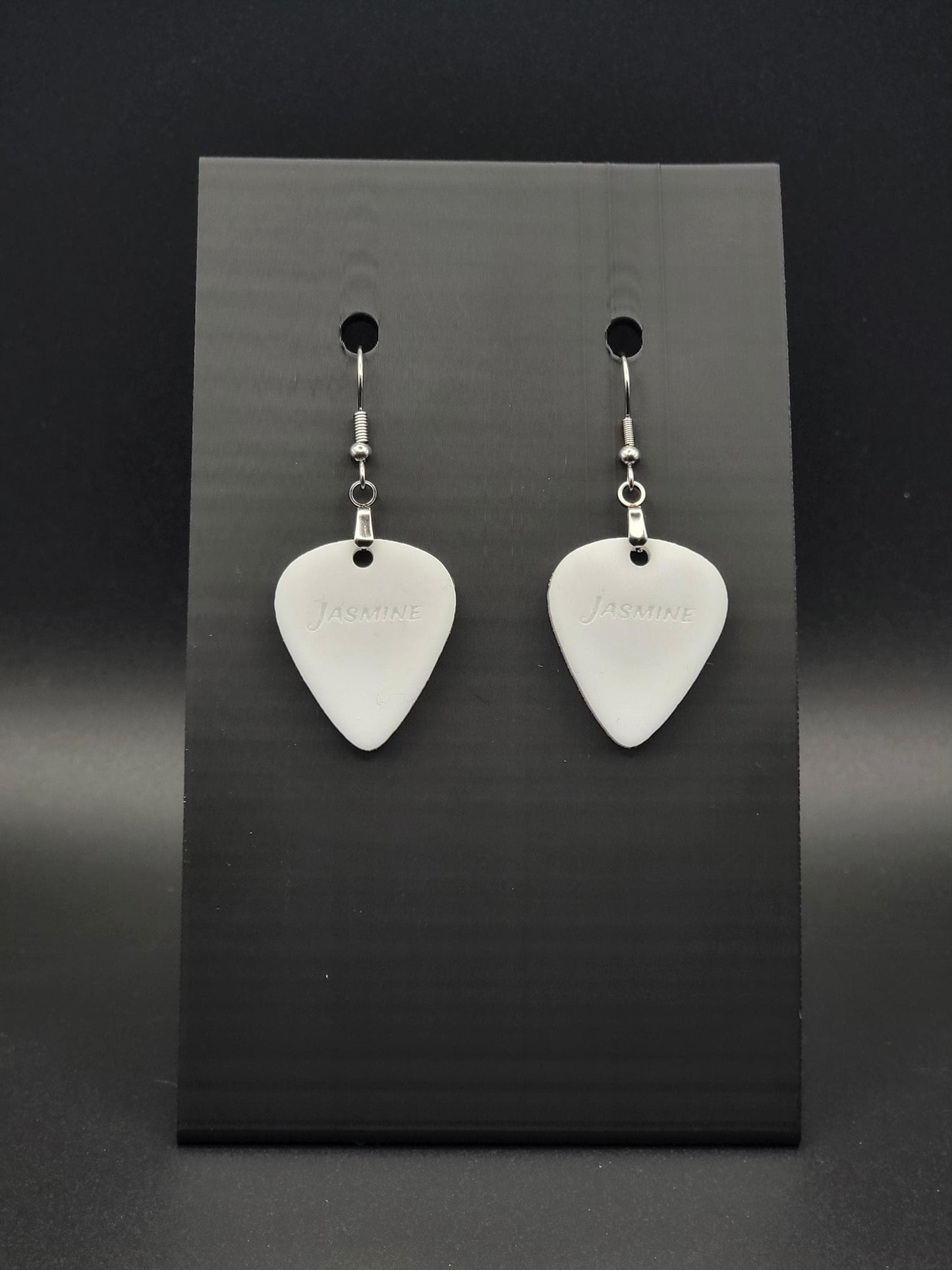 White acrylic guitar pick earring with "Jasmine" engraved, shown on a black jewelry card. Fun, personalized jewelry for music lovers.