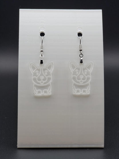 Pair of clear acrylic corgi earrings engraved with a playful face. Shown on a white earring card.