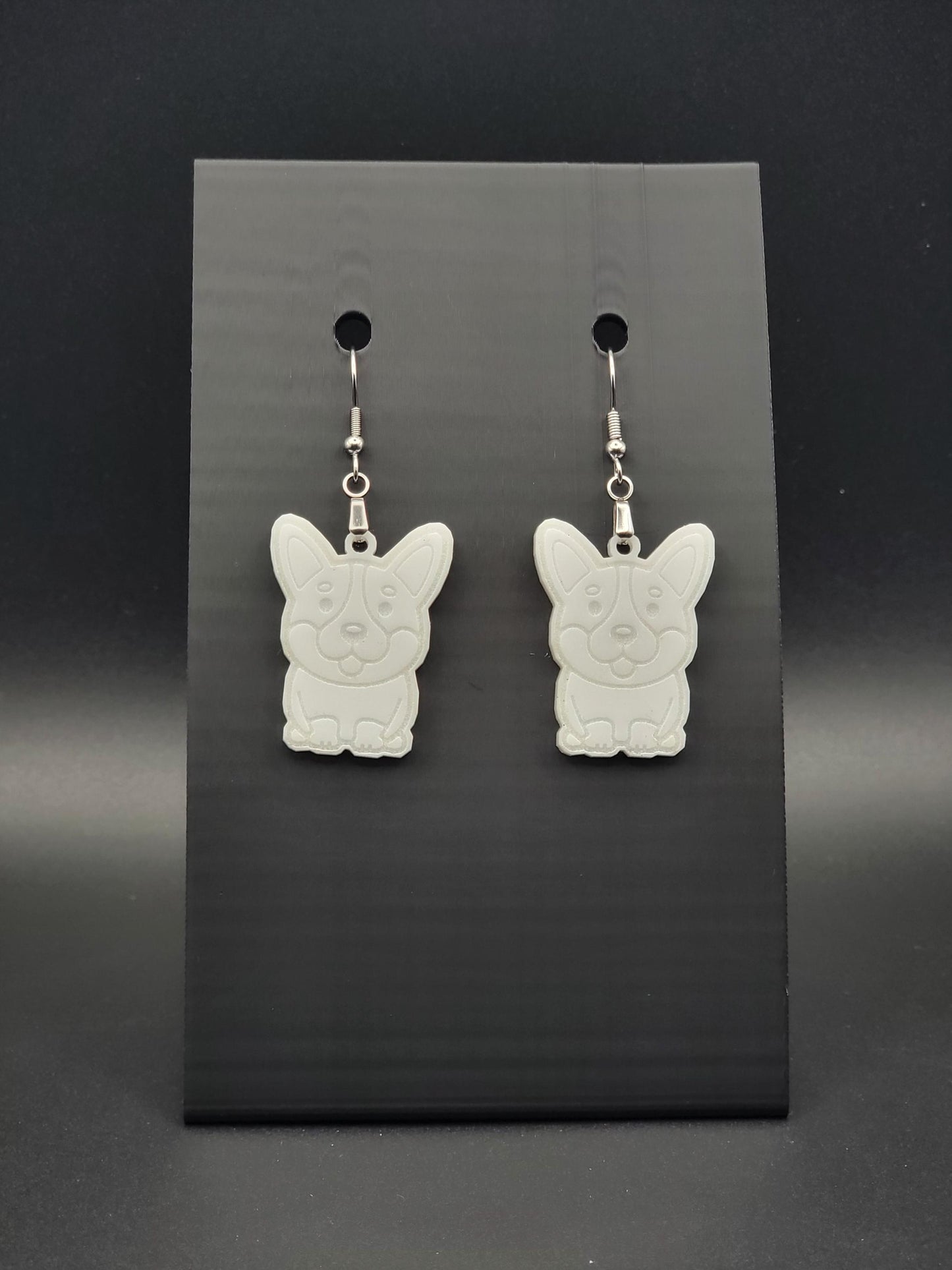Pair of white acrylic corgi earrings engraved with a playful face. Shown on a black earring card.