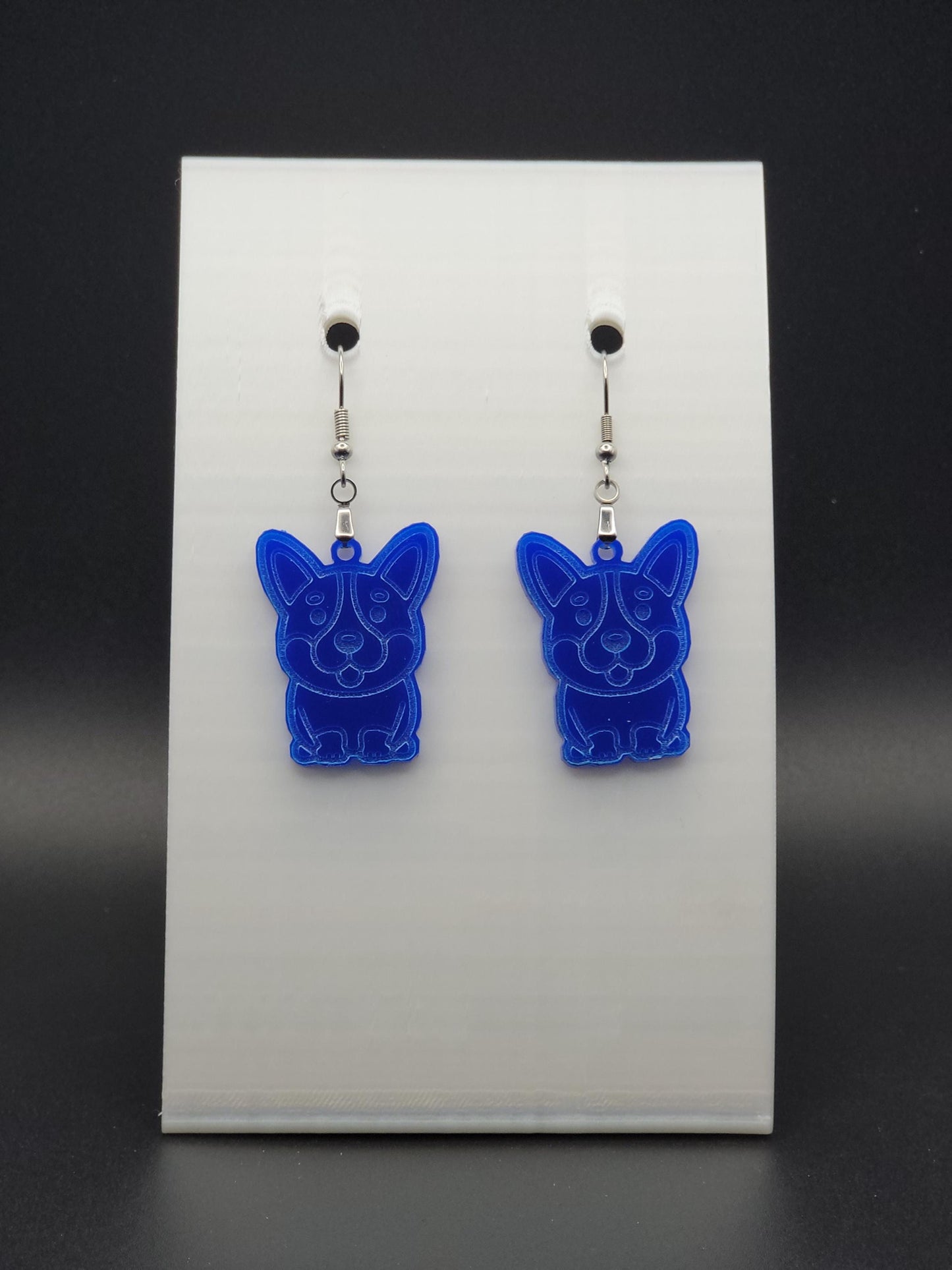 Pair of blue acrylic corgi earrings engraved with a playful face. Shown on a black earring card.