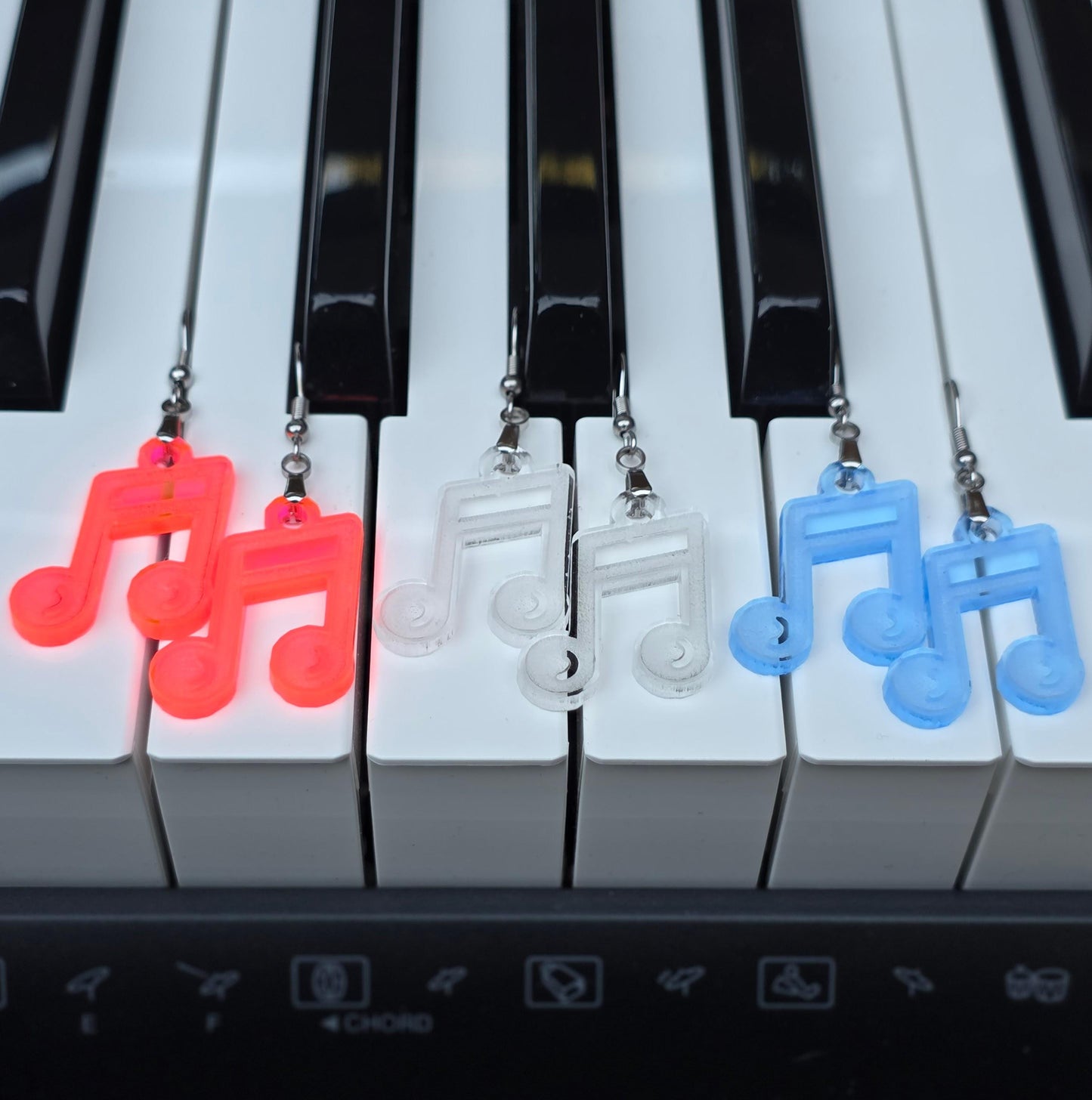 Fluorescent, clear, and clear blue acrylic music note earrings lined up horizontally on the white keys of a piano keyboard. Earrings appear in pairs, organized left to right by color.