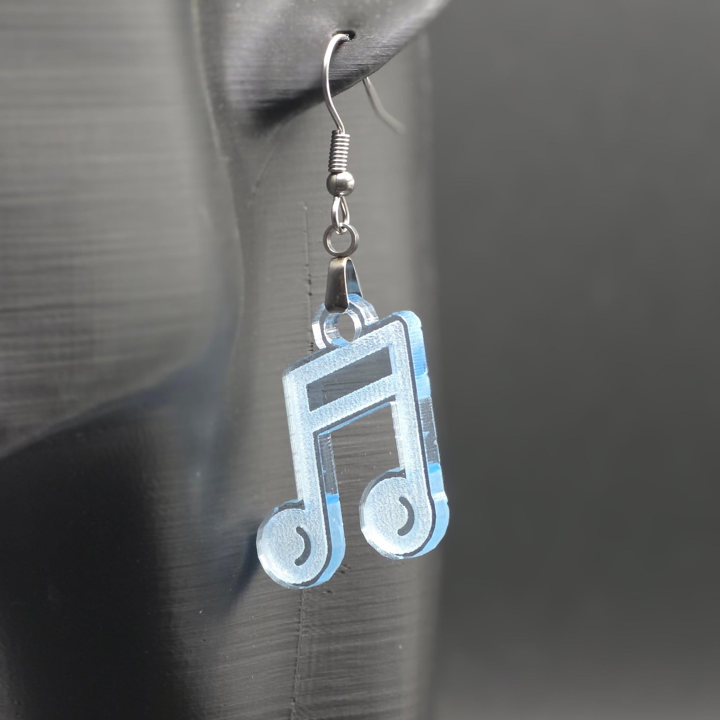 Single earring made of clear blue acrylic shaped like a beamed music note, hanging from a mannequin ear. Matte black background emphasizes color and shine.