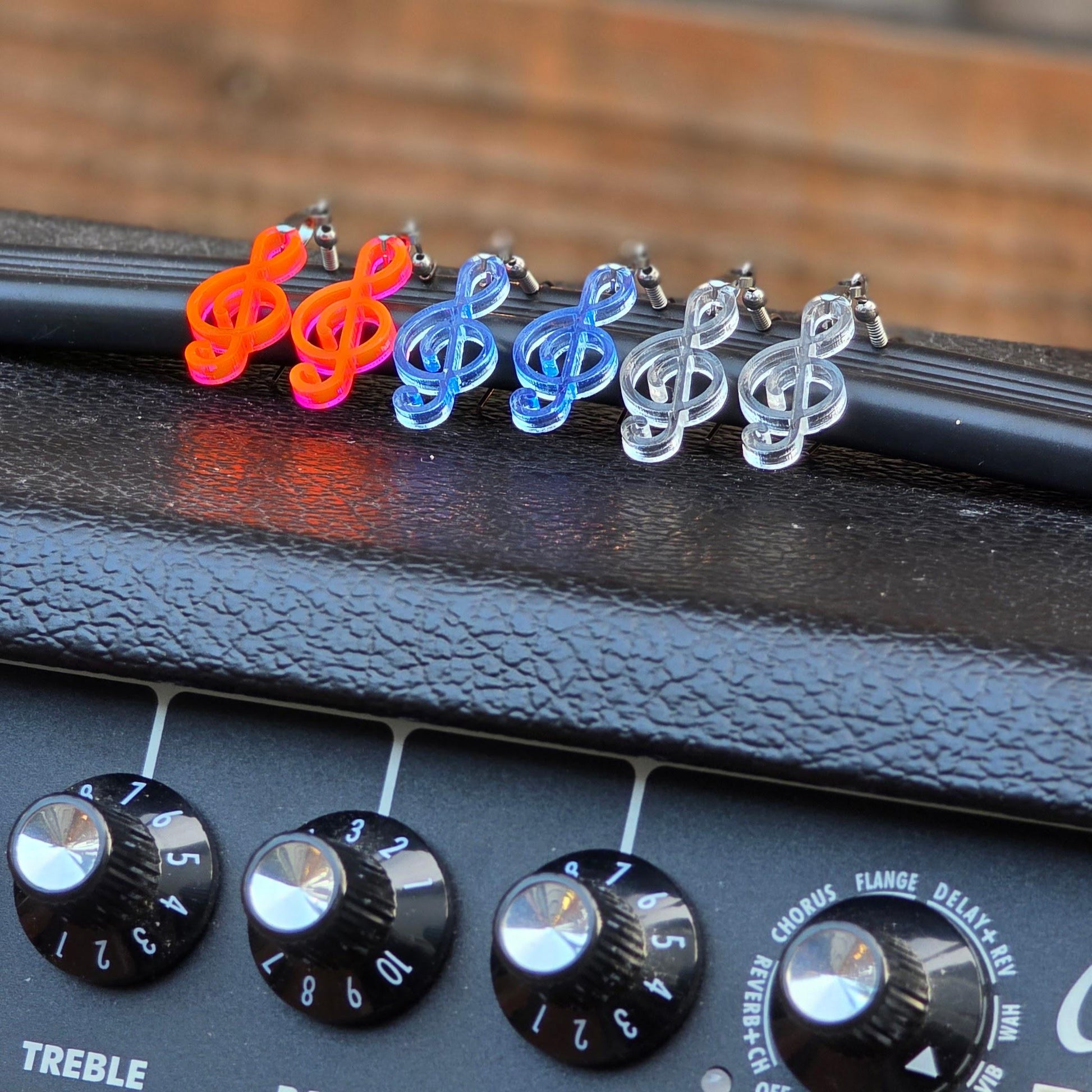 Colorful acrylic treble clef earrings displayed on a guitar amplifier with treble knobs in view. Features fluorescent, clear blue, and clear earrings with stainless steel hooks.
