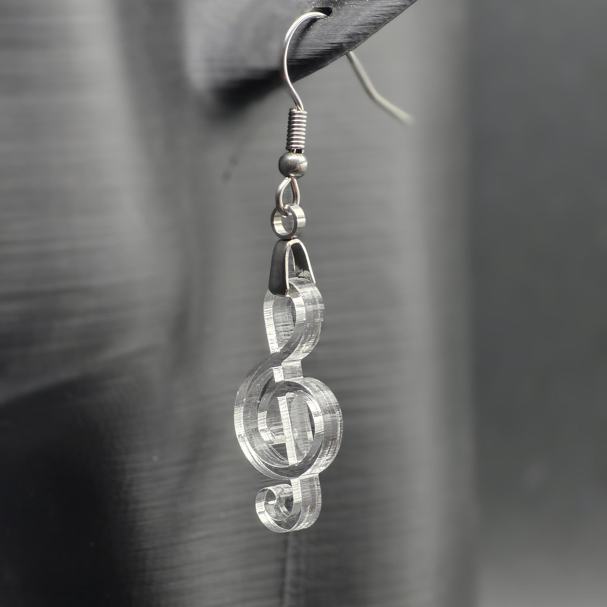 Clear acrylic treble clef earring on matte black mannequin ear, showcasing laser-cut detail and stainless steel hardware.