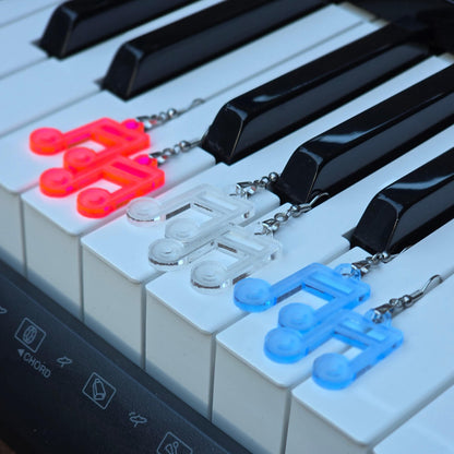 Three pairs of acrylic music note earrings, fluorescent, clear, and clear blue, resting on the white keys of a digital piano. Earrings are spaced out diagonally, emphasizing color variation.