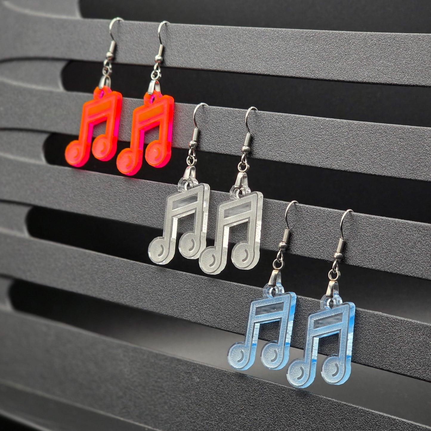 Six acrylic music note earrings in fluorescent, clear, and clear blue displayed in pairs hanging from a gray slatted music stand. Earrings are organized by color in rows with clean black backdrop.