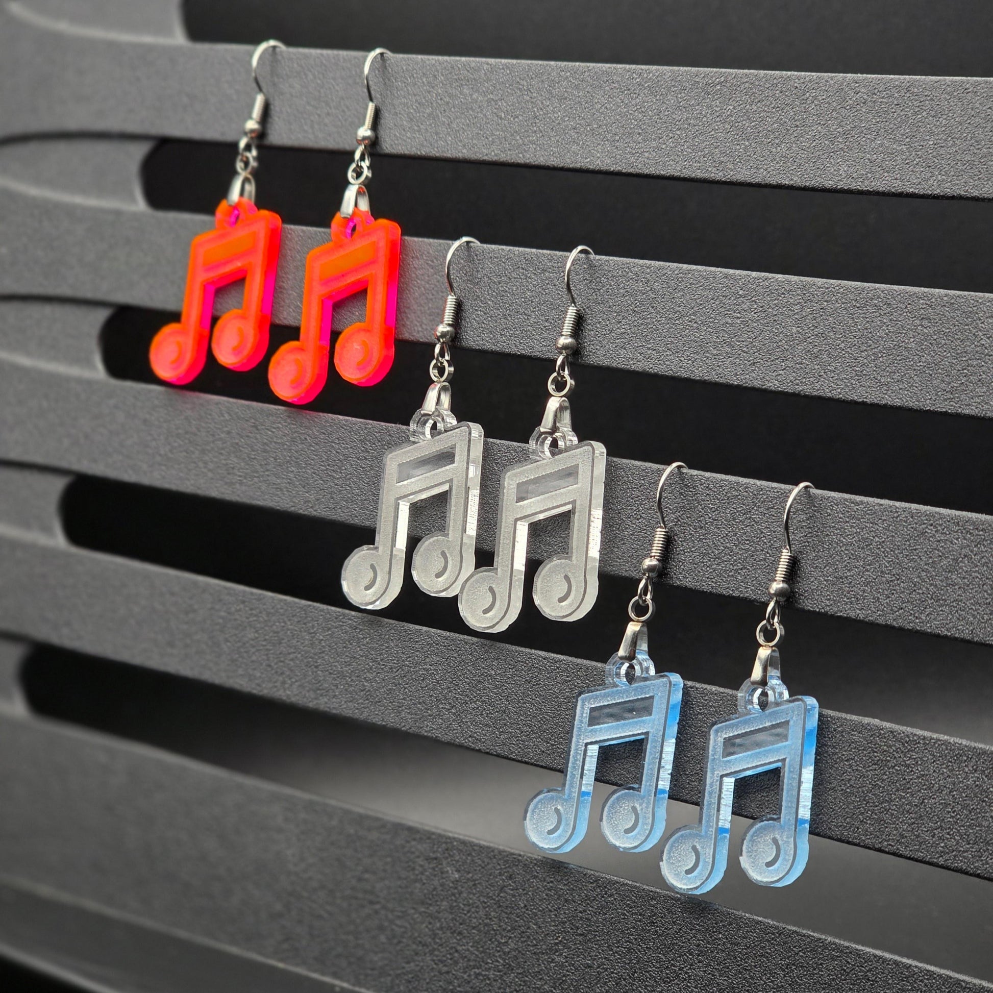 Six acrylic music note earrings in fluorescent, clear, and clear blue displayed in pairs hanging from a gray slatted music stand. Earrings are organized by color in rows with clean black backdrop.